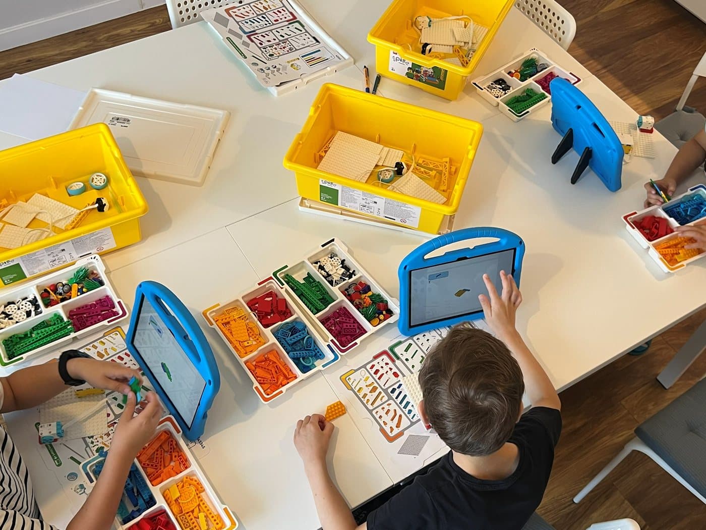 Widok z góry na stół z zestawami LEGO® Education SPIKE Essential i tabletami, dzieci składają modele - w kadrze widać ich dłonie przy pracy